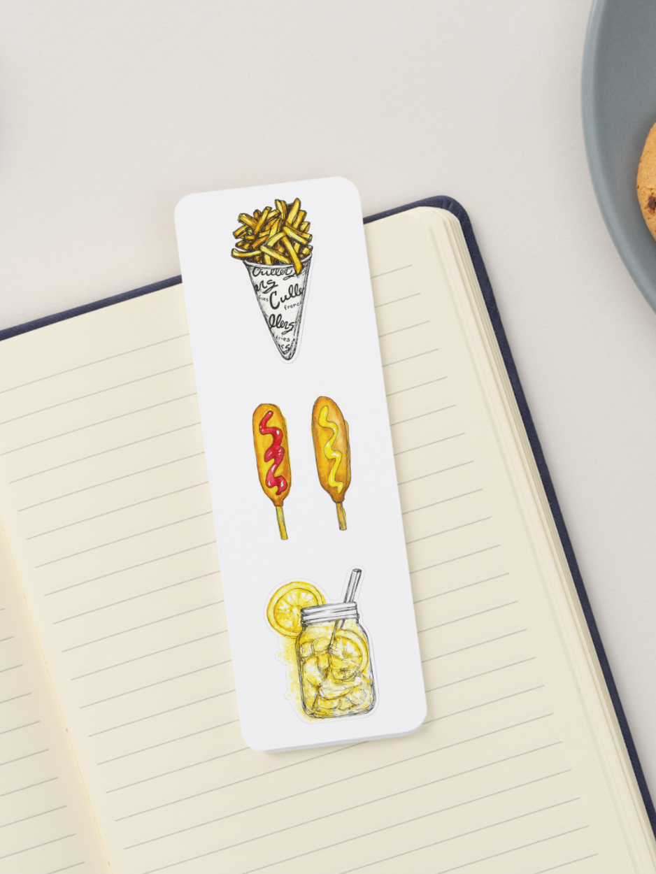 A white rectangular bookmark with illustrated fair food graphics, placed on an open lined notebook. The bookmark shows a cone of french fries, two corn dogs, and a jar of lemon shake-up.