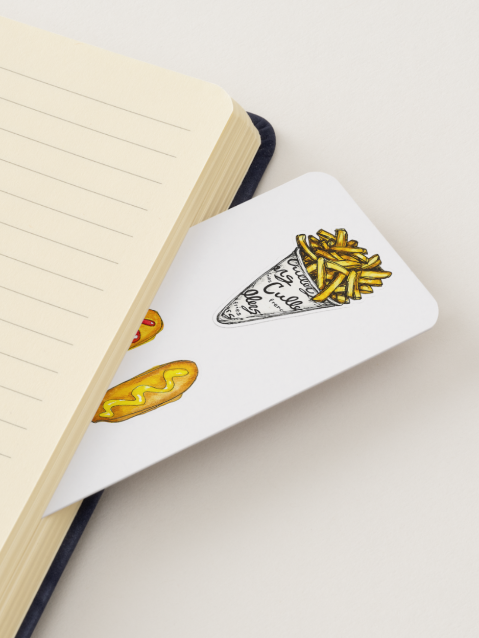 A white rectangular bookmark with a hand-drawn illustration of a french fries cone and a corn dog dog, placed on top of an open cream-colored lined notebook.