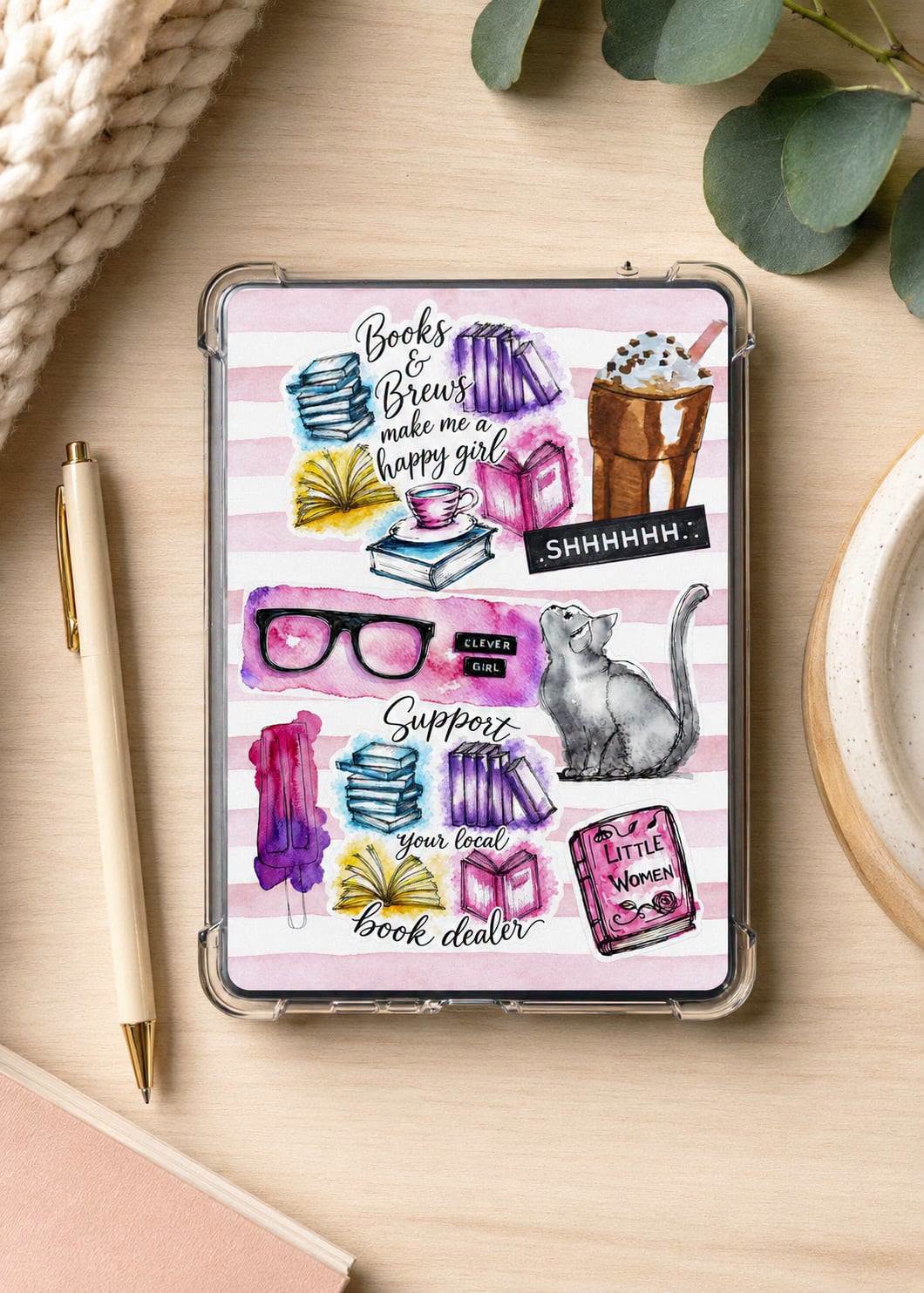 A clear-edged tablet case with a pink striped background and colorful book-themed illustrations, shown on a light wood desk with a pen, knit fabric, eucalyptus leaves, and a plate.